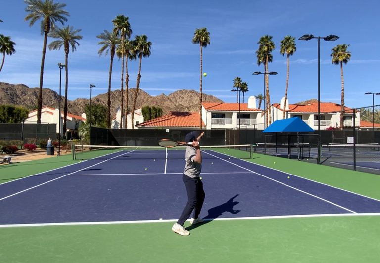 Recreational tennis player serving with a waiter's tray position, racket face open to the sky beside the shoulder instead of dropping behind the back